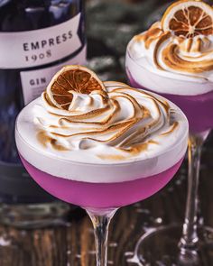 two glasses filled with purple liquid and topped with whipped cream, an orange slice on top