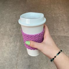 a hand holding a coffee cup with a purple knitted sleeve and green nail polish