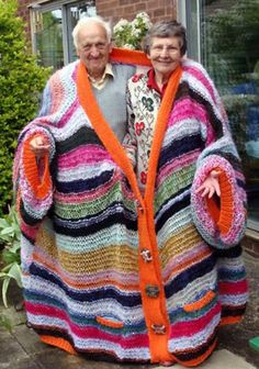 an older couple standing next to each other in front of a house wearing colorful knitted sweaters