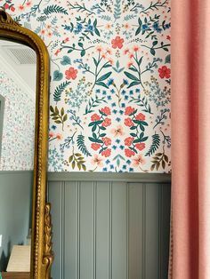 a mirror sitting next to a wall with flowers on it and a pink curtain in front of it