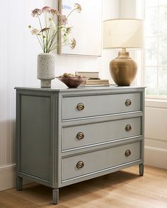 a gray dresser with flowers and a lamp on it in front of a large window