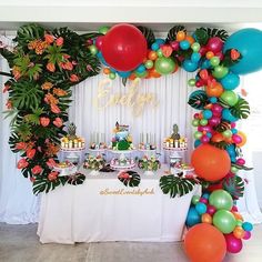 a table topped with lots of colorful balloons and cake next to a wall covered in greenery