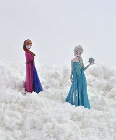 two figurines are standing in the snow, one is holding a flower and the other is wearing a blue dress