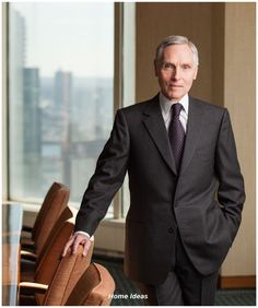 a man in a suit and tie standing next to a chair with his hand on the back of it