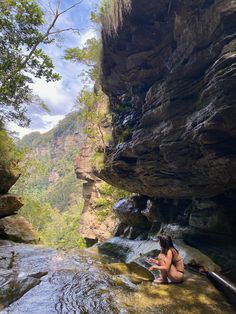 a woman sitting on top of a rock next to a river under a cliff side