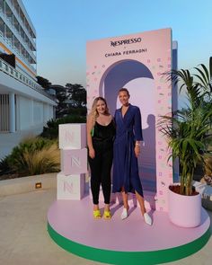 two women standing in front of a sign with the word n on it and palm trees behind them