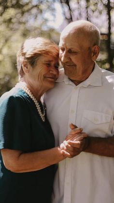 an older man and woman embracing each other in front of some trees with their hands together