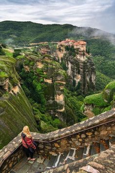 a woman sitting on top of a stone staircase next to a lush green hillside covered in trees