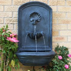 a water fountain in front of a brick wall with pink flowers around it and green leaves on the top