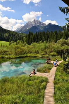 people are sitting at the edge of a small lake with blue water and mountains in the background