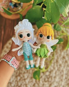 two tiny knitted fairy dolls are held by someone's hand in front of a potted plant