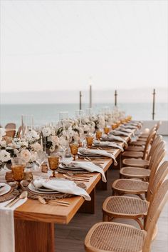 a long table set up with white flowers and place settings for an outdoor dinner by the ocean