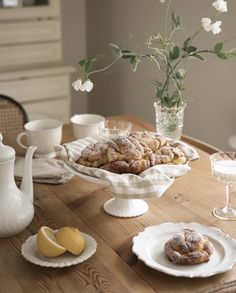 a wooden table topped with white plates filled with pastries and lemon wedges next to a vase full of flowers