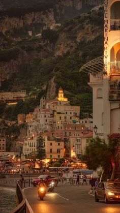 cars are driving down the road in front of some buildings and cliffs at night time