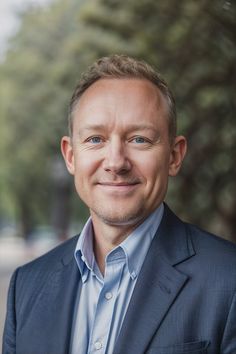 a man wearing a suit and blue shirt smiling at the camera with trees in the background