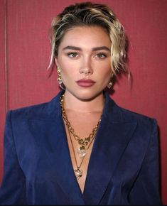 a woman with blonde hair wearing a blue blazer and gold necklace, standing in front of a red wall