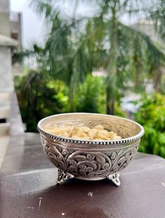 a silver bowl sitting on top of a wooden table next to some trees and bushes