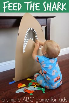 a child playing with a paper shark on the floor next to a cardboard board that says feed the shark