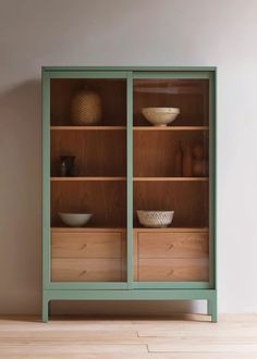 a green bookcase with two glass doors and bowls on the top shelf, in front of a white wall