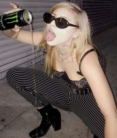 a woman in black and white striped pants with her tongue out, holding a can of monster energy drink