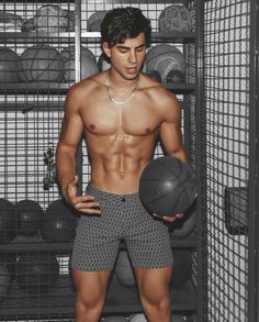 a shirtless young man holding a basketball in front of a rack with other sports balls