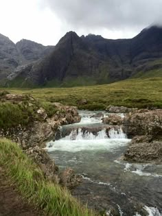 a river running through a lush green valley surrounded by mountains in the distance with grass and rocks on either side