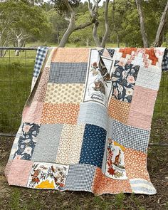 a patchwork quilt is hanging on a fence in the grass with trees behind it