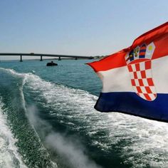 the flag of croatia is flying from a boat in the water with a bridge in the background