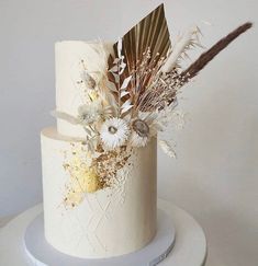 a white and gold wedding cake decorated with feathers, dried flowers and leaves on top