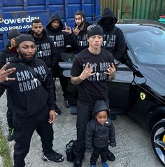 a group of young men standing next to a black car with their hands in the air