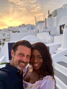 a man and woman posing for a photo in front of white buildings with the sun setting behind them