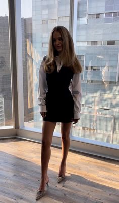 a woman standing in front of a window wearing high heels and a white shirt dress