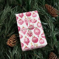 a present wrapped in pink and white paper with christmas ornaments on it, surrounded by pine cones