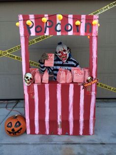 a costumed clown sitting in front of a carnival booth with candy boxes and pumpkins