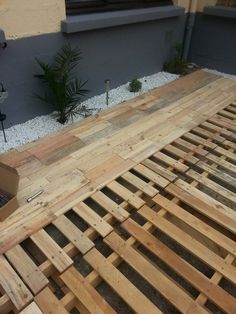 an unfinished wooden bed frame sitting in front of a gray building with gravel on the ground