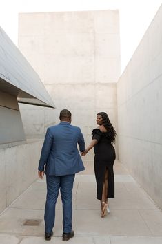 a man and woman holding hands walking down a sidewalk in front of a building with concrete walls