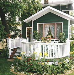 a small green and white house with flowers in the front yard, next to a deck