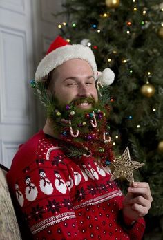 a man with a beard wearing a christmas sweater and holding an ornament in front of a christmas tree