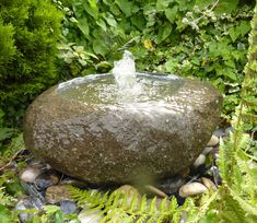 a water fountain in the middle of some plants and rocks with green foliage around it