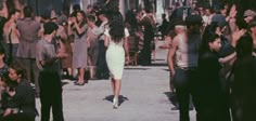 a woman in a white dress walking through a crowd of people with their hands up