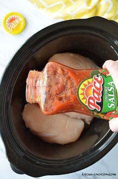 a hand holding a jar of hot sauce in a slow cooker filled with chicken