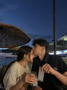 two people sitting at a table with drinks in front of an umbrella on the beach