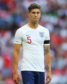 a man with tattoos standing in front of a soccer field wearing a white shirt and blue shorts