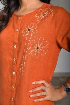 a woman wearing an orange shirt with white flowers on the front and side, standing in front of a wall