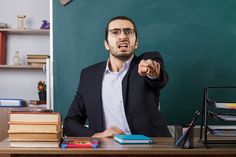 a man sitting at a desk in front of a blackboard pointing to the side