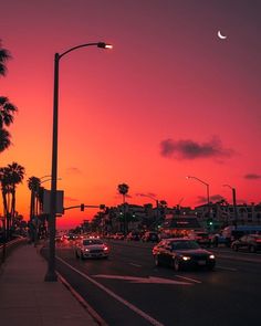 cars are driving down the street at sunset with palm trees in the foreground and a half moon