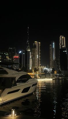 a white boat is docked in the water near some tall buildings at night with its lights on