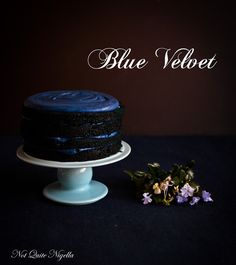 a blue velvet cake sitting on top of a white cake stand next to purple flowers