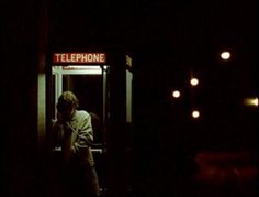 a man standing in front of a telephone booth at night with his cell phone up to his ear