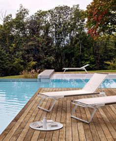 two lounge chairs sitting on top of a wooden deck next to a swimming pool with trees in the background